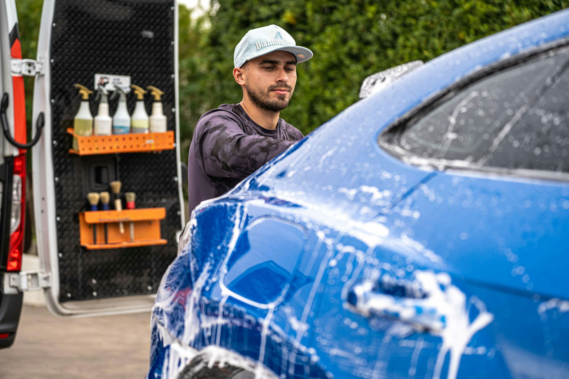 Detailer myjacy niebieskie auto obok regalu z chemia detailingowa